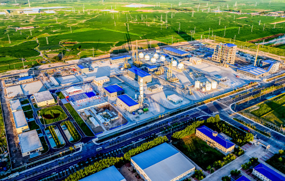 This image shows an aerial view of an industrial complex or factory. The facility has multiple buildings, storage tanks, and infrastructure typical of a manufacturing or processing plant. Surrounding the complex are green fields and wind turbines, indicating a rural or semi-rural location with a focus on renewable energy nearby. The shadows suggest the photo was taken in the late afternoon or early morning. If you need specific information or analysis about this image, please let me know!