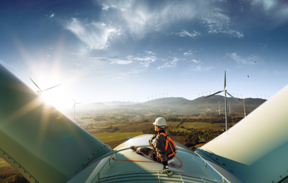 The image shows a person sitting on top of a wind turbine, wearing safety gear including a helmet and a harness. The person appears to be inspecting or maintaining the turbine. The background features a landscape with multiple wind turbines scattered across the fields and hills, under a clear blue sky with some clouds. The scene suggests a focus on renewable energy and the maintenance of wind power infrastructure.