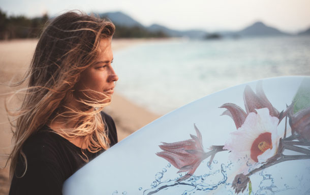 Person with long hair holding a surfboard with floral and water design on a beach with mountains in the background.