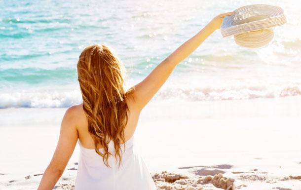 The image shows a woman sitting on a sandy beach facing the ocean. She has long, wavy hair and is wearing a white sleeveless top or dress. She is holding a wide-brimmed hat in her right hand, raised above her head. The scene is bright and sunny, with the ocean waves gently rolling in the background.