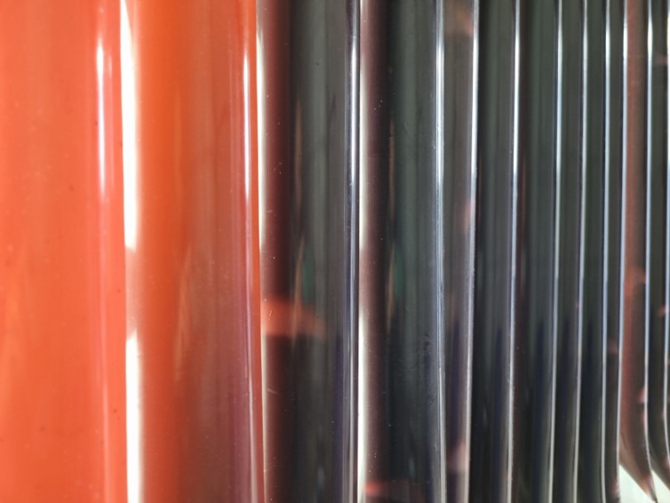 The image shows a close-up view of a series of vertical, cylindrical tubes or rods. The left side features tubes with a reddish-orange color, while the right side has tubes with a reflective, metallic, or dark surface. The tubes are arranged closely together, creating a pattern of alternating colors and reflections. The surface of the tubes appears smooth and shiny. This image could be part of an industrial or mechanical component, such as heat exchanger tubes or pipes.