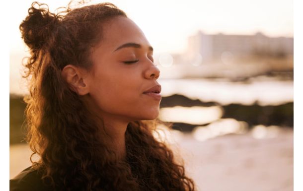 The image shows a person with curly hair styled in a half-up bun. The background features a beach setting with a blurred view of the ocean and a building in the distance, suggesting a serene and possibly sunset or sunrise scene.
