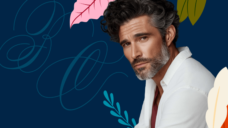 The image shows a stylish middle-aged man with dark curly hair and a salt-and-pepper beard, wearing a white shirt over a dark red top. He’s posed against a dark blue decorative background with abstract floral and leaf elements, giving it a polished, editorial-style look.