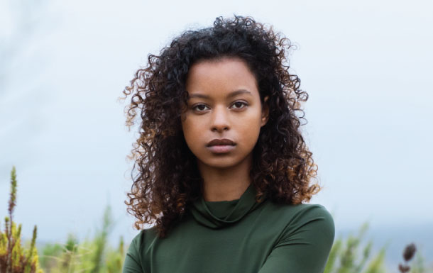 The person in the image has curly hair and is wearing a dark green long-sleeve top. The background appears to be outdoors with some greenery and a cloudy sky. If you need any specific information or analysis about the image, please let me know!