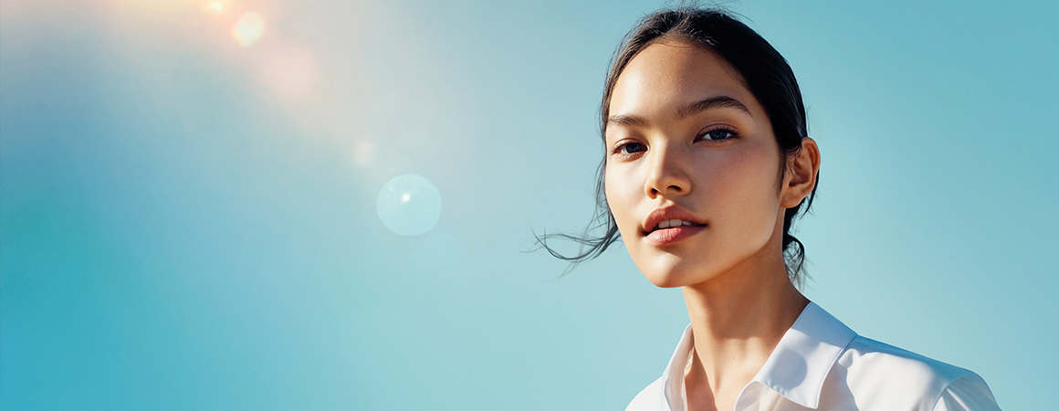 Person wearing a white shirt standing outdoors against a clear blue sky with sunlight and lens flare.