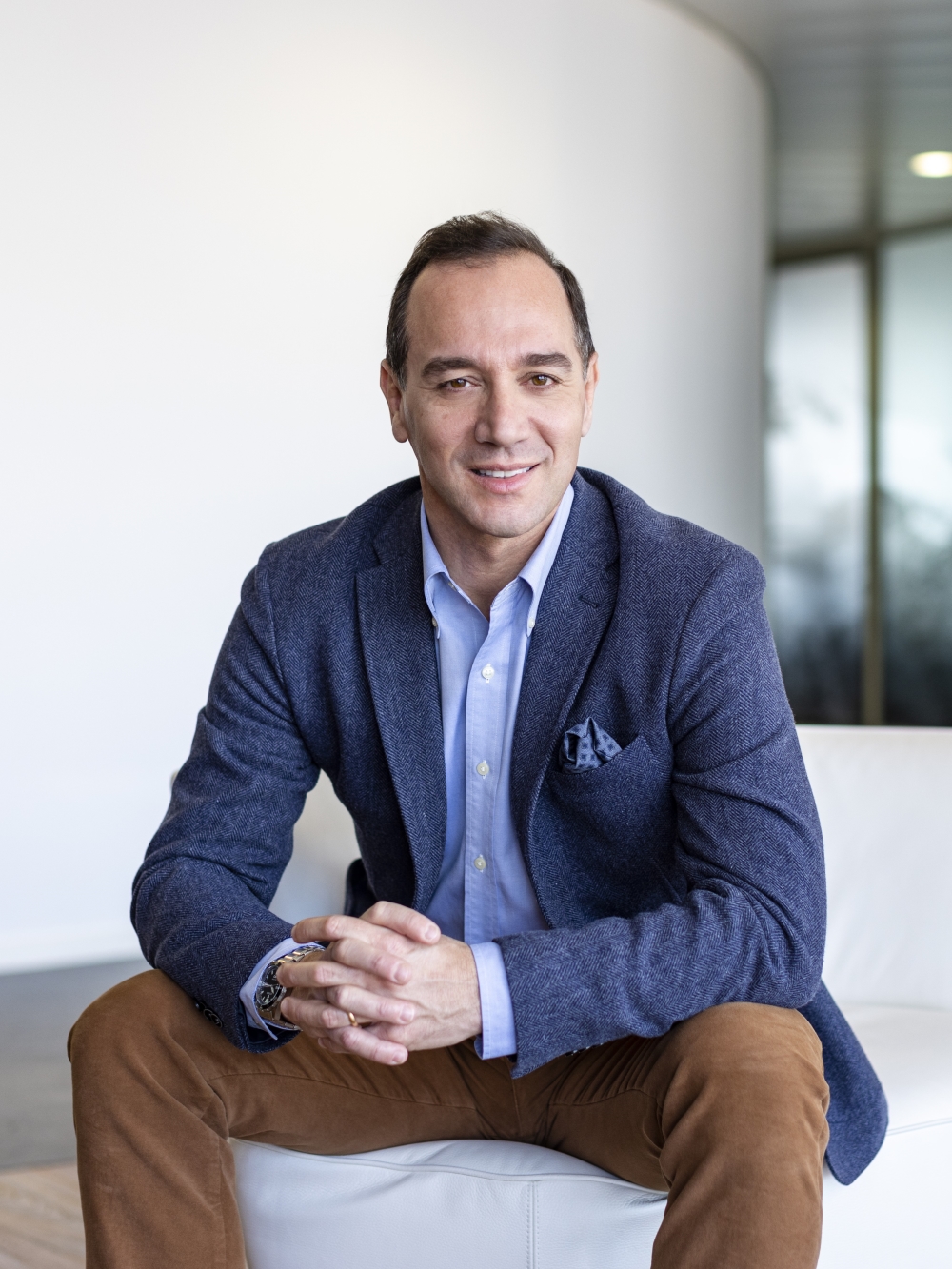 The person in the image is wearing a blue blazer over a light blue button-up shirt. They have paired this with brown pants. The individual is seated on a white piece of furniture with their hands clasped together. The background appears to be an indoor setting with a curved white wall and a window. The overall attire suggests a smart-casual or business-casual style.