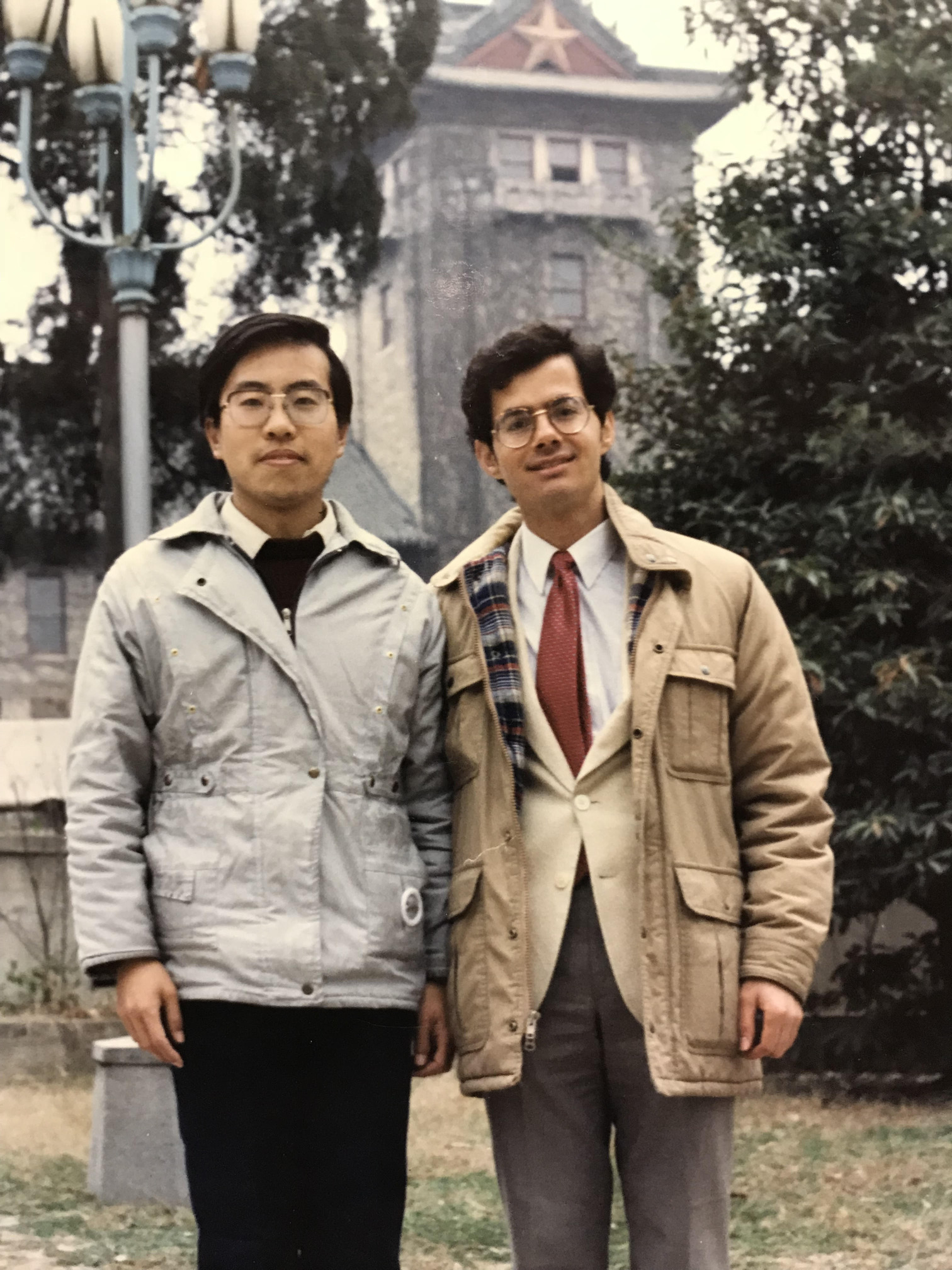 The image shows two men standing outdoors in front of a building with a star on its roof. Both are wearing jackets suitable for cool weather. The man on the left is dressed in a light gray jacket over a dark sweater and white shirt, while the man on the right is wearing a beige jacket over a light-colored blazer, white shirt, and red tie. There is greenery around them, including trees and bushes, and a lamp post is visible on the left side of the image. The overall setting appears to be a park or garden area.