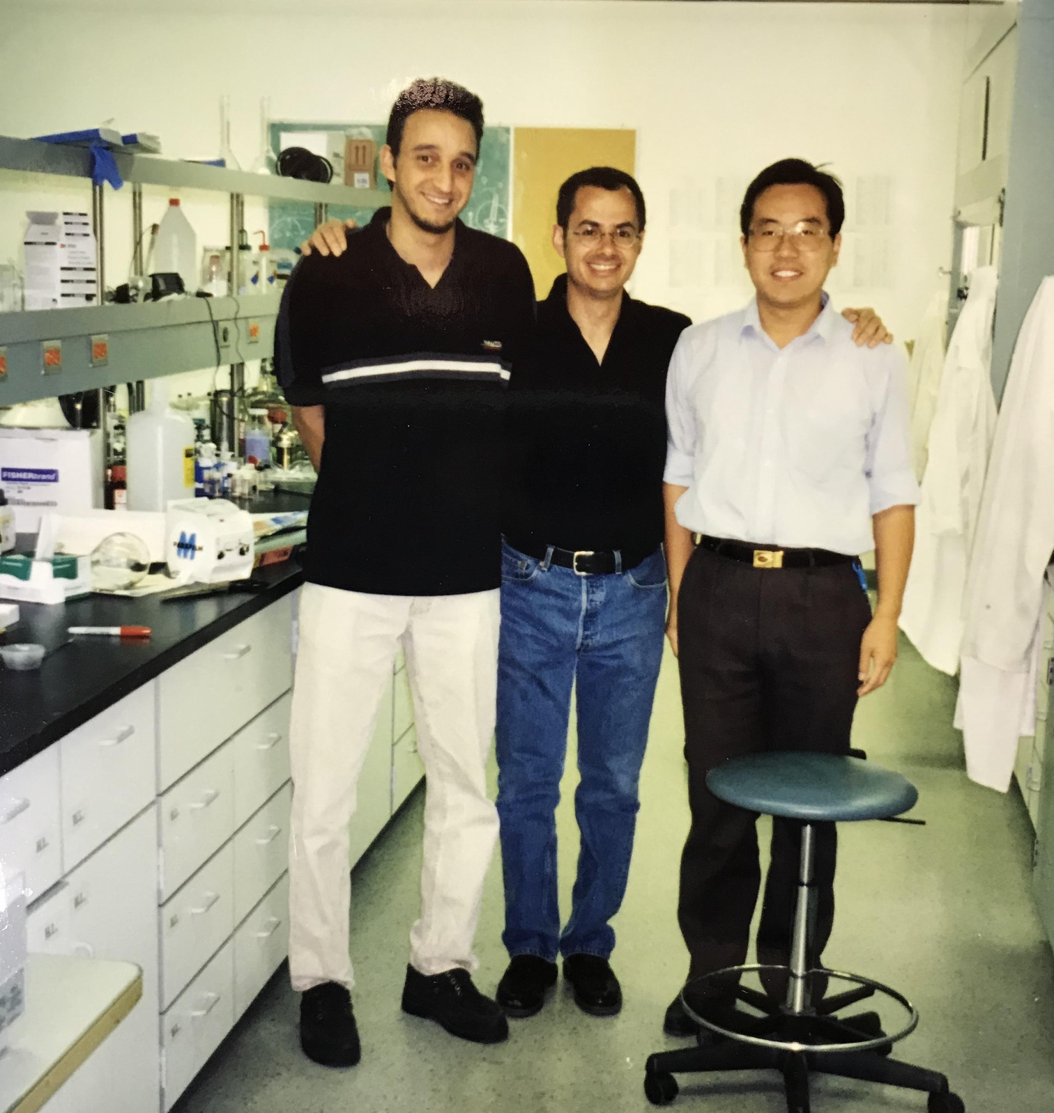 The image shows three men standing in a laboratory setting. The man on the left is wearing a black shirt with white and gray stripes and light-colored pants. The man in the middle is dressed in a black shirt and blue jeans. The man on the right is wearing a light blue shirt with the sleeves rolled up and dark pants. They are standing close to each other, with the man on the left and the man on the right each placing a hand on the shoulders of the man in the middle. The laboratory has various scientific equipment and supplies on the counters, and there is a stool with a blue seat in front of the men. The room appears well-lit and organized.