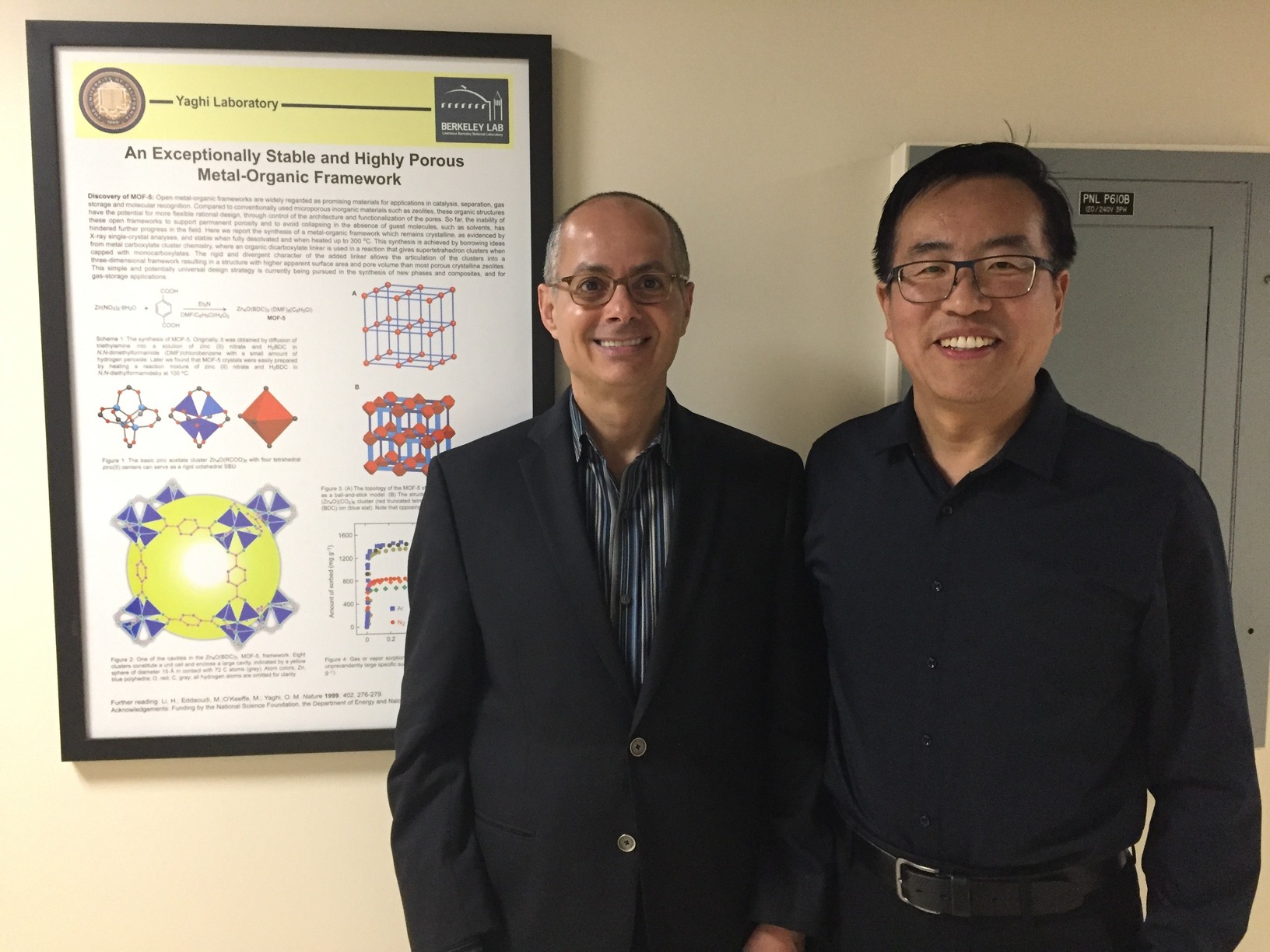 The image shows two men standing side by side indoors. The man on the left is wearing a dark suit jacket over a striped shirt, while the man on the right is dressed in a dark button-up shirt tucked into dark pants with a belt. Behind them, there is a framed scientific poster titled "An Exceptionally Stable and Highly Porous Metal-Organic Framework" from the Yaghi Laboratory at Berkeley Lab. The poster includes diagrams and text related to the research topic. To the right of the men, there is a gray electrical panel labeled "PNL P6108." The setting appears to be a professional or academic environment, likely related to scientific research.