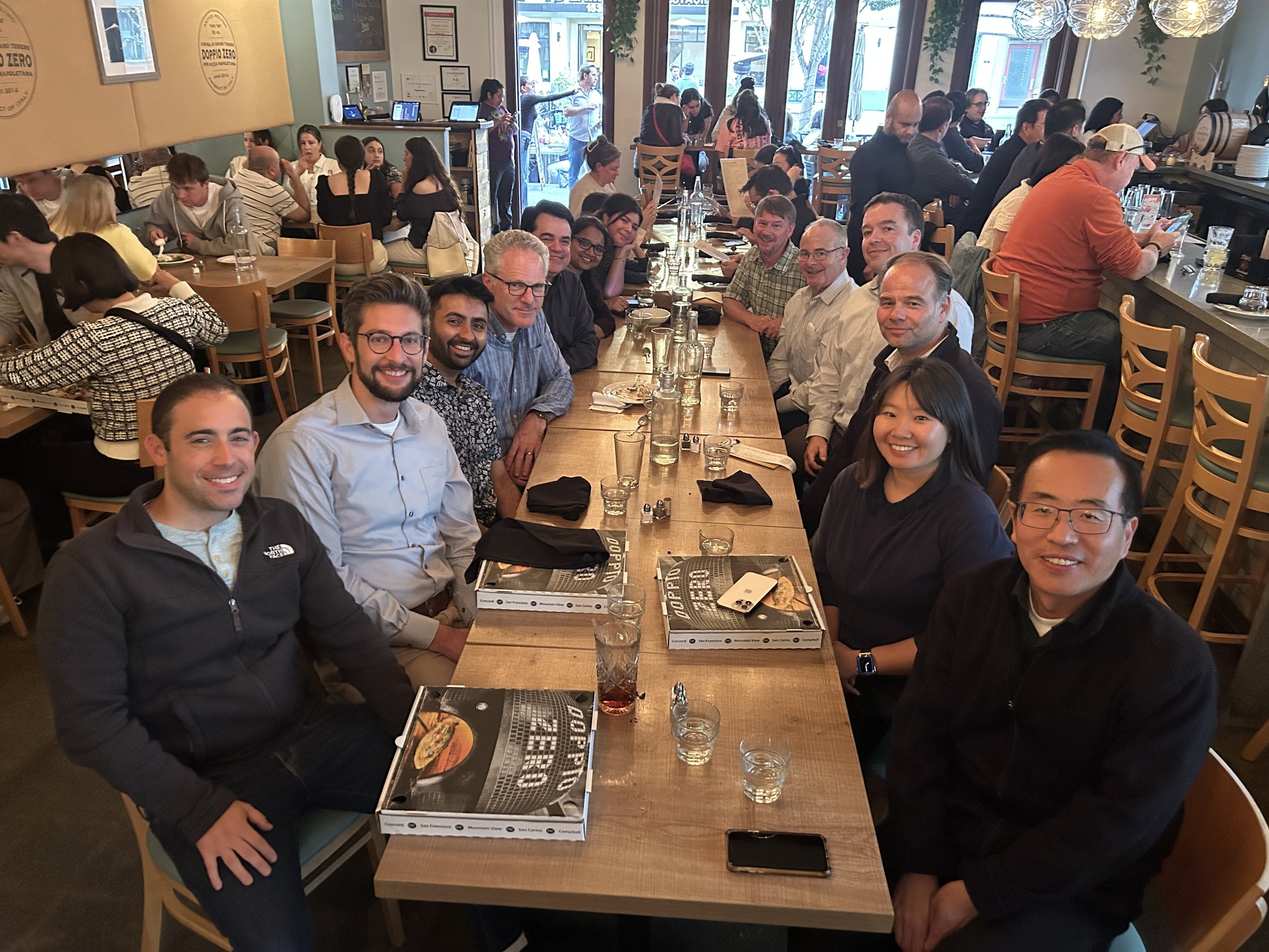 The image shows a large group of people seated around a long wooden table in a restaurant. The table is set with glasses, napkins, and menus or pizza boxes labeled "DOPPIO ZERO." The restaurant appears busy with many patrons seated at other tables and the bar area. The atmosphere looks lively and social. The decor includes hanging lights and some greenery. The people at the table seem to be gathered for a meal or a social event.