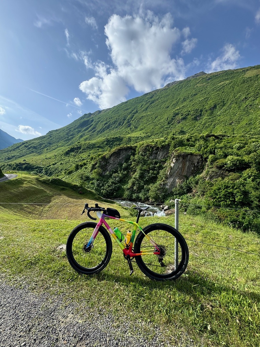 The image shows a colorful road bicycle parked on a grassy area with a scenic backdrop of green hills and a partly cloudy blue sky. The bike has a vibrant frame with colors including pink, yellow, green, and red. There are two water bottles attached to the frame, one green and one yellow. The setting appears to be a peaceful, natural outdoor location, possibly a mountainous or hilly region. The bike is positioned near a gravel path or road.