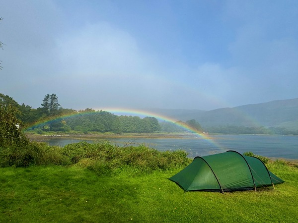 The image shows a green tent set up on a grassy area near a body of water, likely a lake or river. In the background, there is a forested area with trees and hills. A vibrant rainbow arcs across the sky, and a faint secondary rainbow is also visible above it. The sky is mostly clear with some light clouds, creating a serene and picturesque outdoor camping scene.