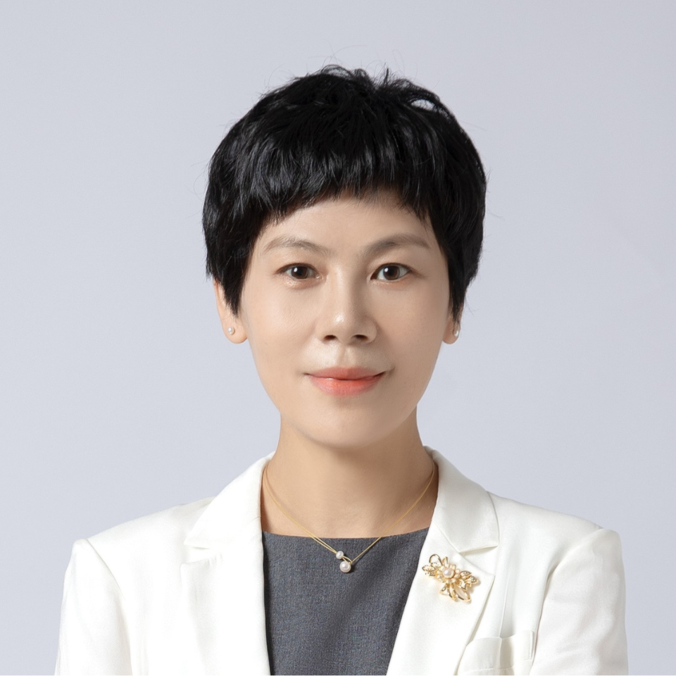 The person in the image is wearing a white blazer over a gray top. They have short, dark hair and are accessorized with a delicate gold necklace featuring a small pendant and a decorative brooch on the left lapel of the blazer. The background is plain and light-colored, which keeps the focus on the individual.